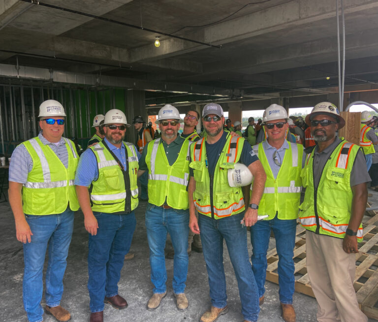 Group of construction workers in hard hats and safety vests posing for a photo on a worksite.
