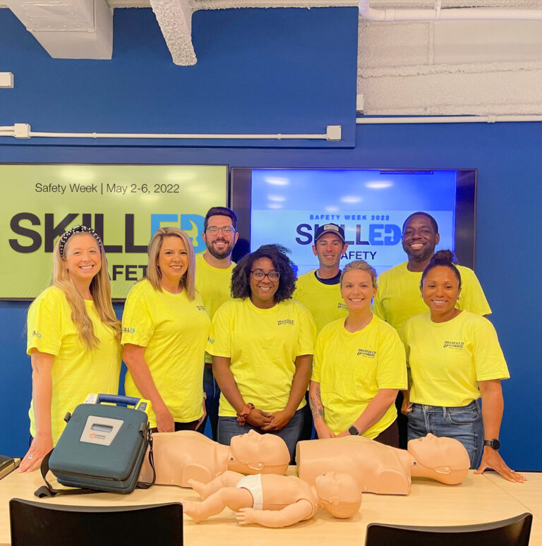 Group of people in yellow shirts posing for safety week event with medical training equipment and a presentation screen in the background.
