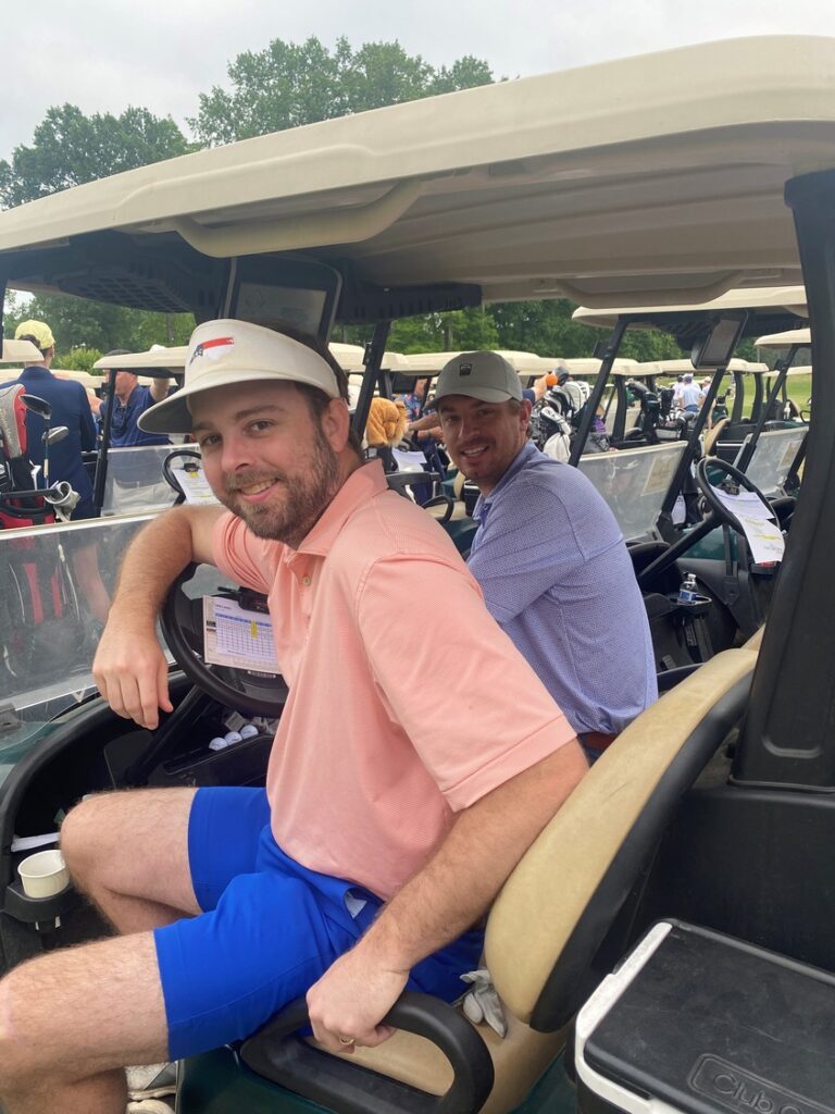 Two men seated in a golf cart, looking at the camera, with others in the background on a golf course in Charlotte, NC.