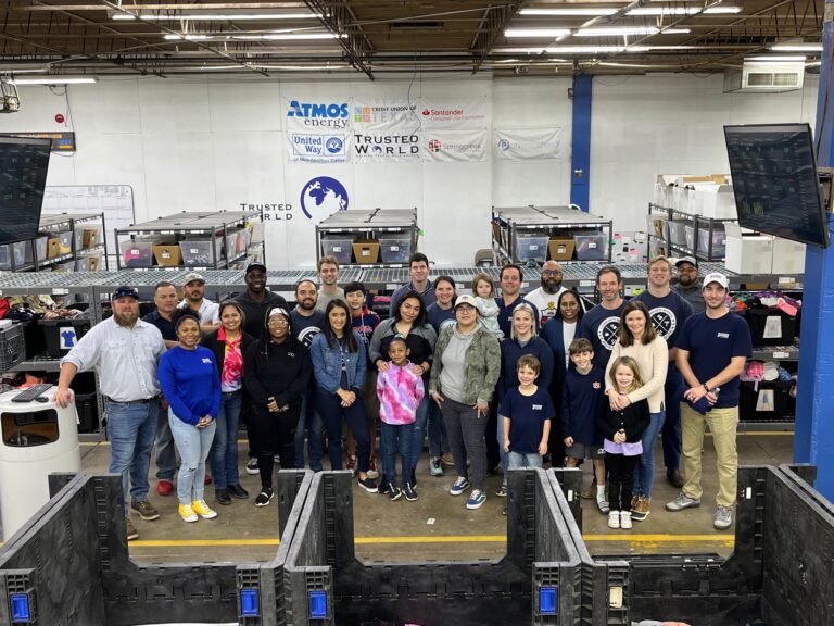 A diverse group of volunteers posing for a photo in a Dallas warehouse with supplies and equipment in the background.