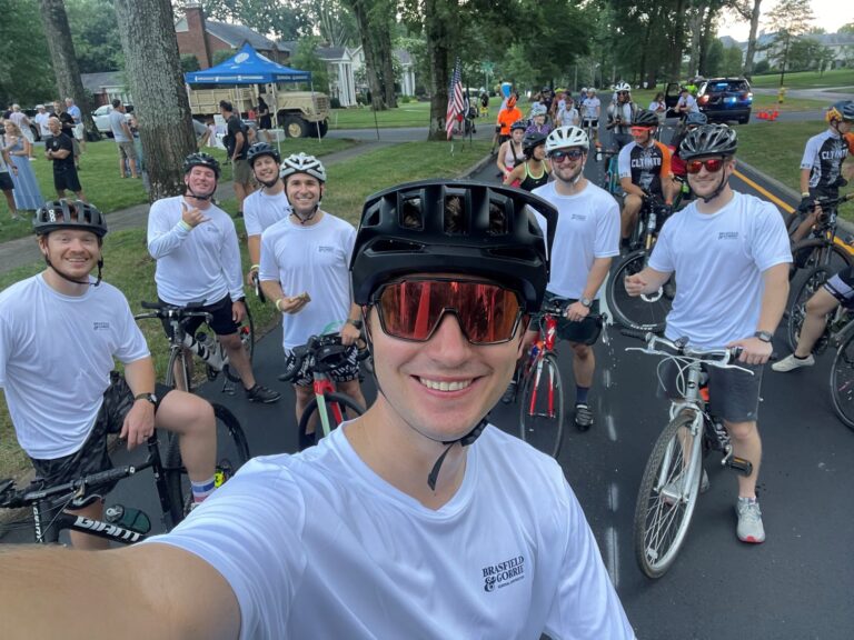 Group of cyclists posing for a selfie before a ride in Charlotte, NC, with spectators and an American flag in the background.