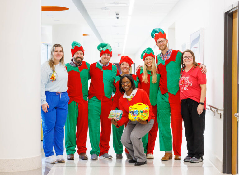 A group of adults dressed in colorful costumes and casual attire posing in a hallway in Jackson, Mississippi, with some wearing elf costumes and one person kneeling in front holding toys.