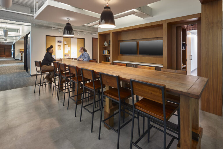 Two individuals having a conversation in a Dallas office break room with a long communal table and pendant lighting.