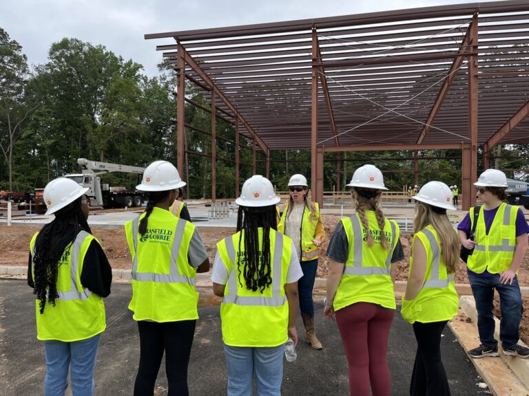 A group of students wearing hard hats and reflective vests stand in front of a metal framework at a Clemson University construction site. A female construction worker speaks to them about the project.