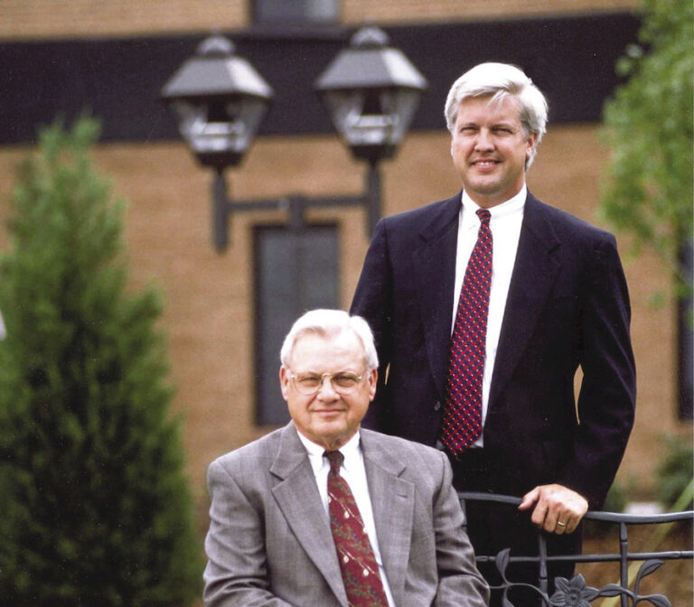 Two men in business suits posing for a photo outdoors, one seated and the other standing behind him.