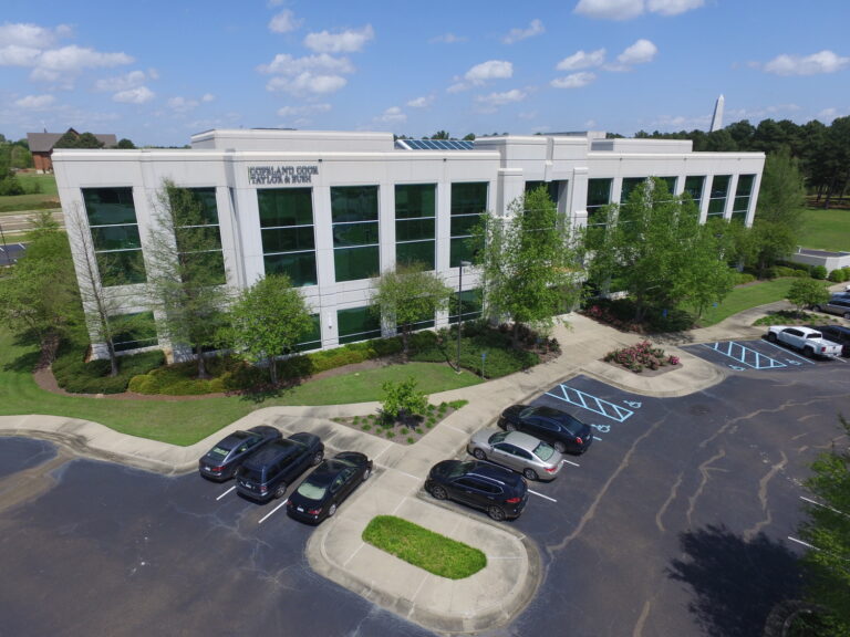 Aerial view of a modern three-story office building with parked cars and surrounding landscaping.