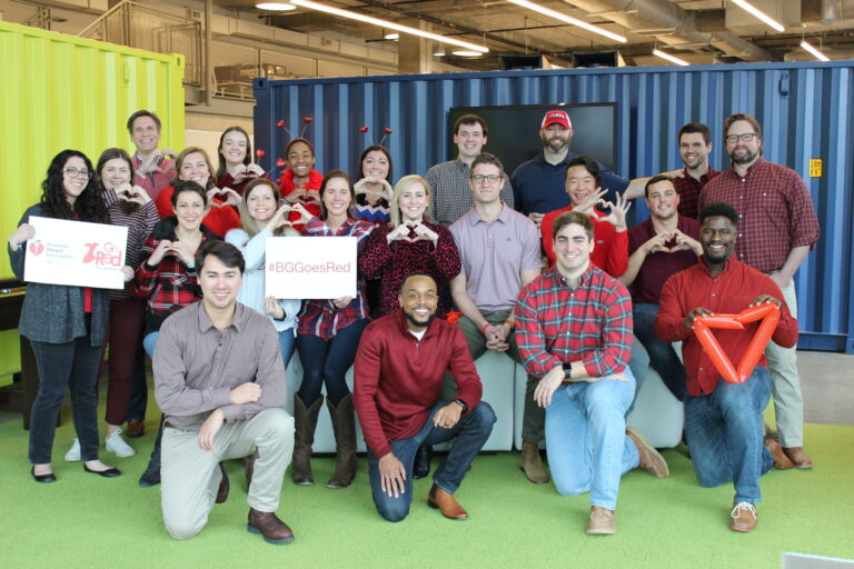 Group of smiling people in a casual office setting, some holding heart-shaped items and wearing red, possibly supporting a heart health awareness campaign.