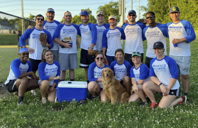 A co-ed softball team posing for a group photo on a field with a dog at the front center.