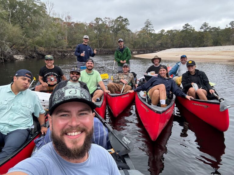 Group of men smiling in three canoes on a river in Jacksonville, Florida, surrounded by trees with a sandy beach in the background.