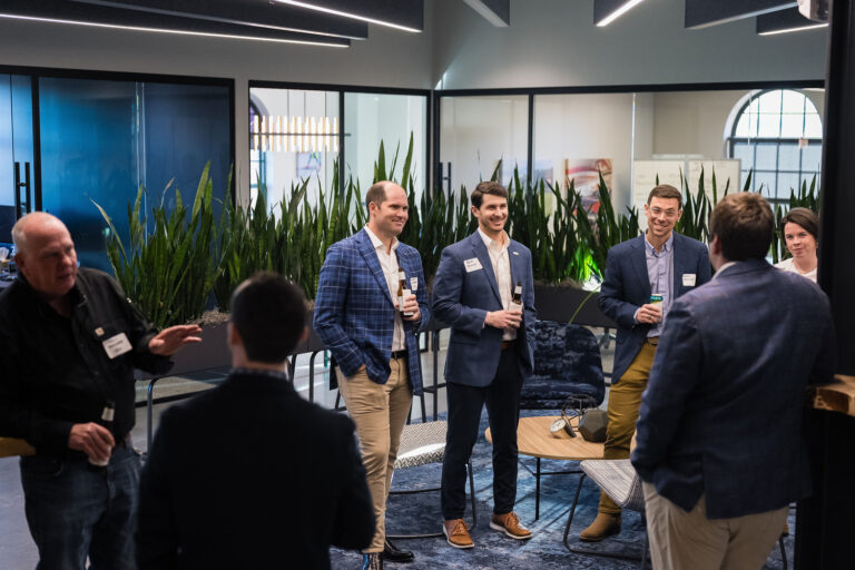 In the Greenville office of Brasfield and Gorrie, a group of people in business attire stand and converse amid plants and modern furniture, creating a lively yet professional atmosphere.