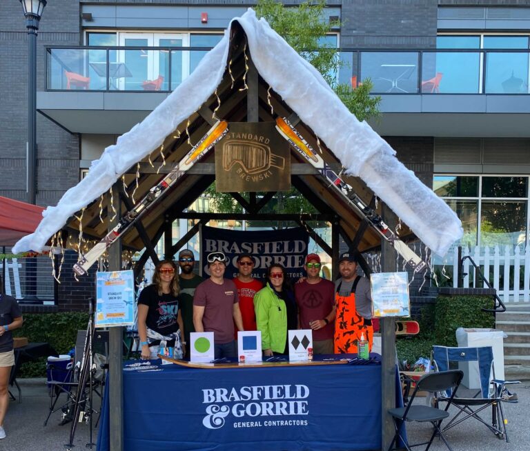 Group of people standing at a promotional booth with signage for brasfield & gorrie, adorned with a mock snowy roof, at an outdoor event.