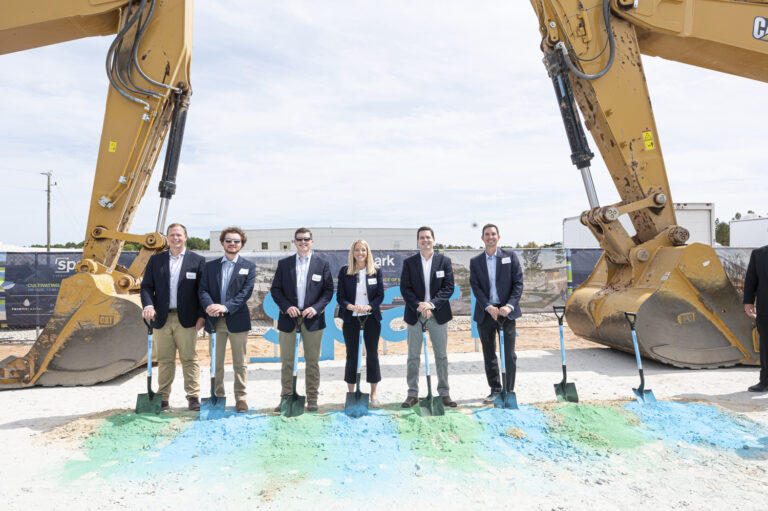 Group of individuals in business attire participating in a groundbreaking ceremony with shovels and construction equipment in the background.
