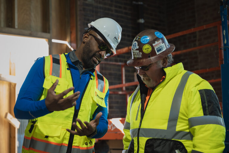 Two construction workers in reflective vests and hard hats are discussing a project on a worksite.