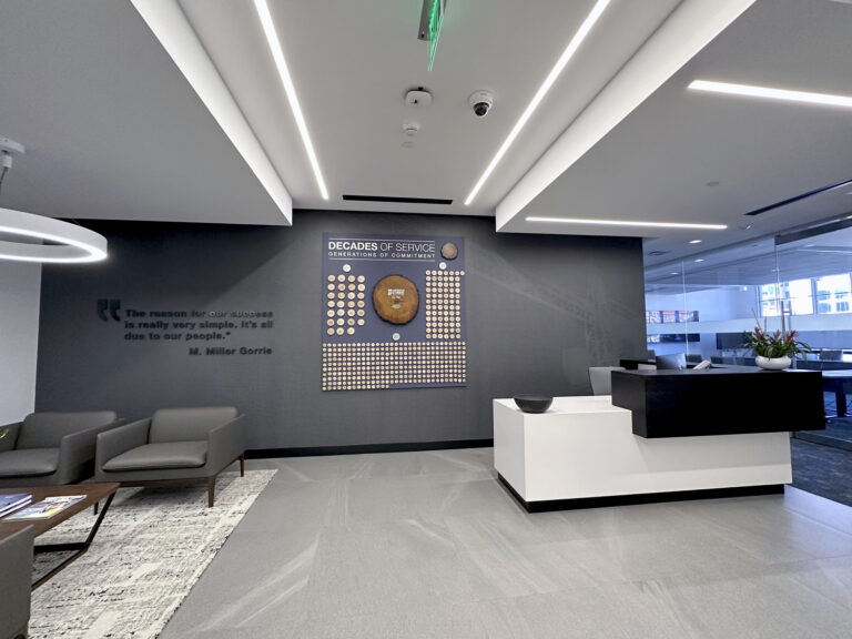 Modern office lobby with a tribute wall celebrating 'decades of service' and a quote, featuring a minimalist reception desk and contemporary lighting.