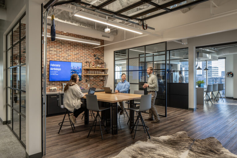 Modern office space in Charlotte, North Carolina, with exposed brick, employees having a meeting at a central table, and a screen displaying 'February birthdays'.