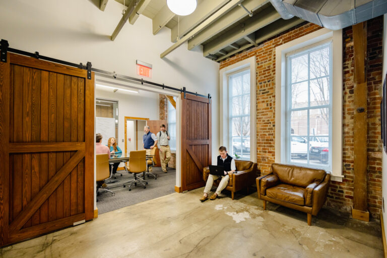 Modern office space with exposed brickwork, large wooden sliding doors, and employees engaged in work and discussion.