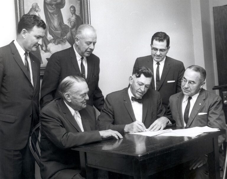 Group of six businessmen in formal attire, one signing a document while others observe.