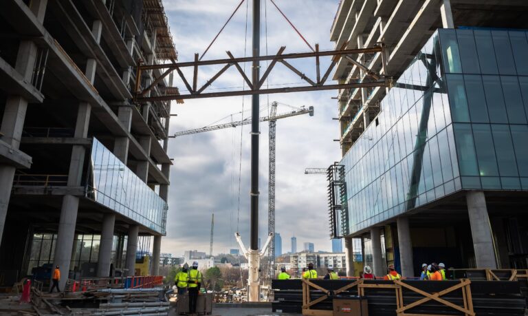 Workers look up at a crane placing a truss on a tall building