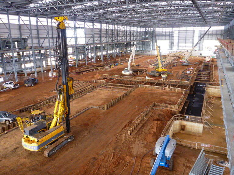 Large indoor construction site with multiple excavators and machinery working on extensive infrastructure excavations under a metal framework.