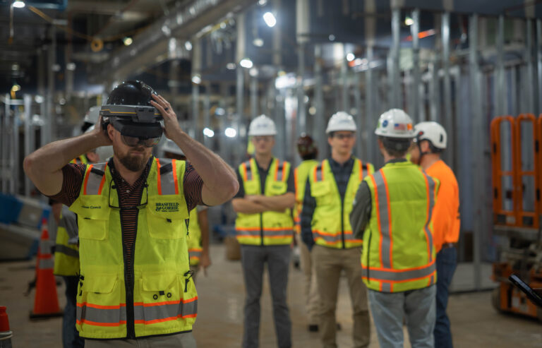 Construction worker using augmented reality headset on site while colleagues discuss in the background.