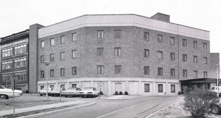 Historical photograph of a brick hospital building with a curved facade on a city street, accompanied by parked cars and a clear sky.