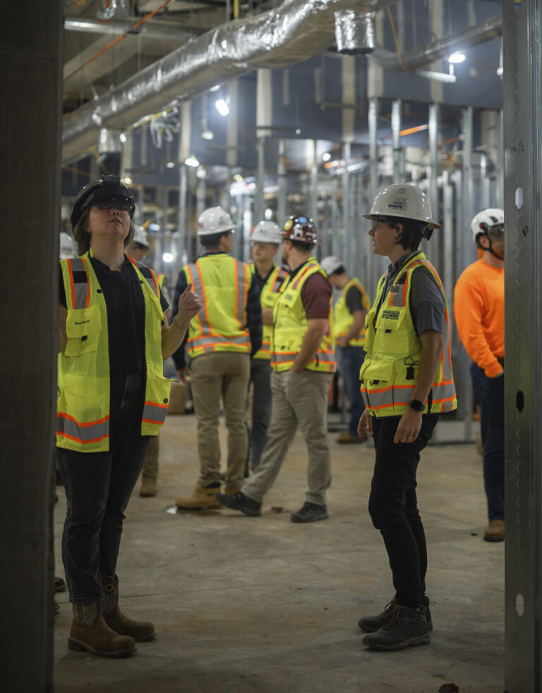 Construction workers in hard hats and reflective vests, harnessing technology, inspecting a site indoors.