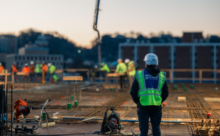 A self-perform construction site worker wearing a helmet and reflective vest oversees other workers amidst building materials during early morning.