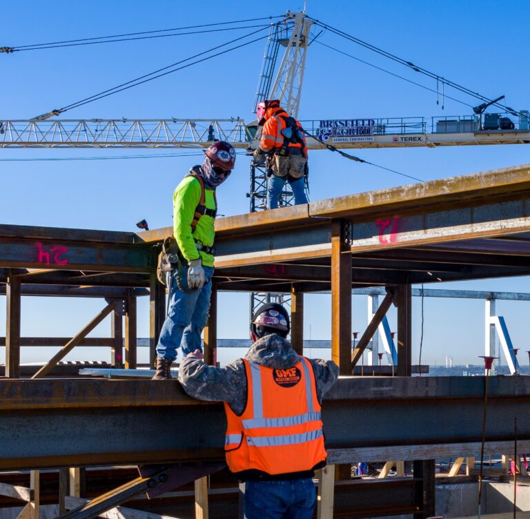 Three trades workers on a steel beam at a building site, with one supervising as two others secure beams under a crane.