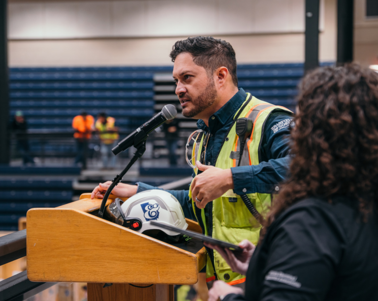 A tradesman in a safety vest and helmet speaks at a podium in a gymnasium, gesturing with one hand, as another person listens.