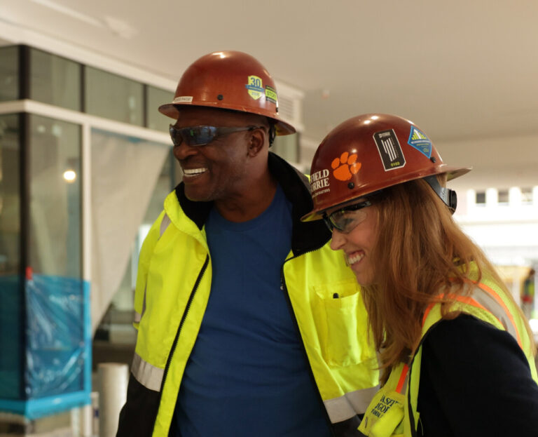 Two construction workers in hard hats and reflective vests share a laugh at a construction site, exemplifying the company's commitment to social responsibility.