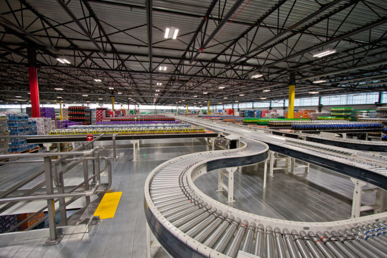 Interior of a large industrial distribution warehouse with extensive conveyor belt systems transporting colorful packaged goods under a metal roof.
