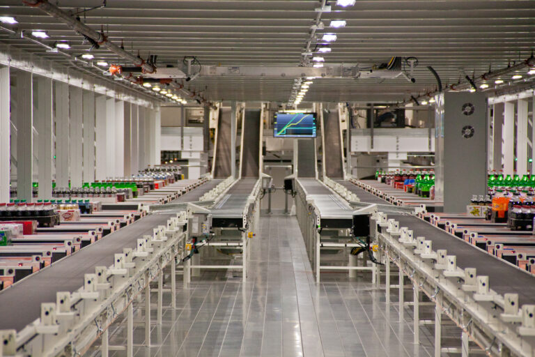 Interior of a modern beverage distribution facility with multiple conveyor belts carrying bottles, and digital displays overhead.