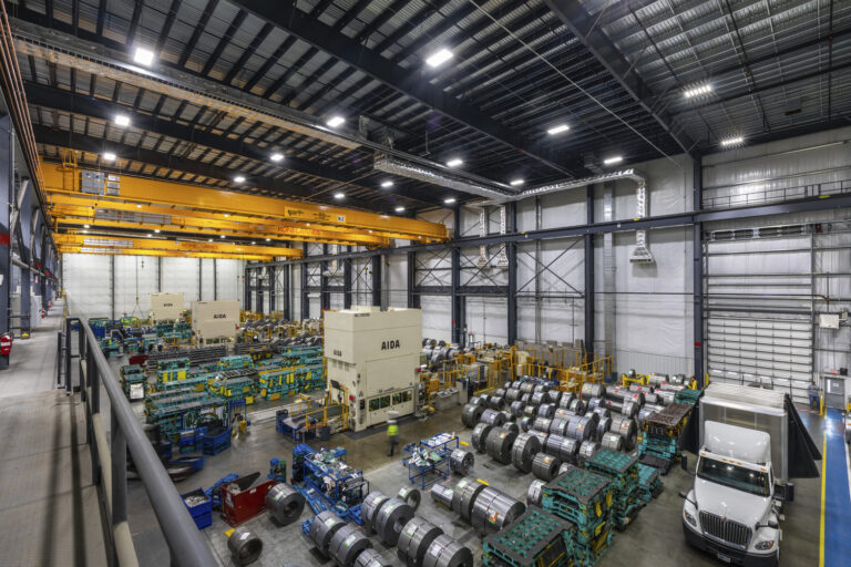 Spacious industrial sector warehouse interior with multiple machinery units, metal coils, and a parked truck, illuminated by ceiling lights.