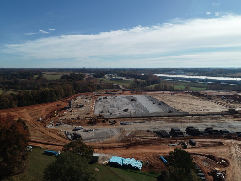 Aerial view of a large construction site with cleared land, building foundations in progress, Auto Draft construction vehicles, and materials, surrounded by a rural landscape and trees.