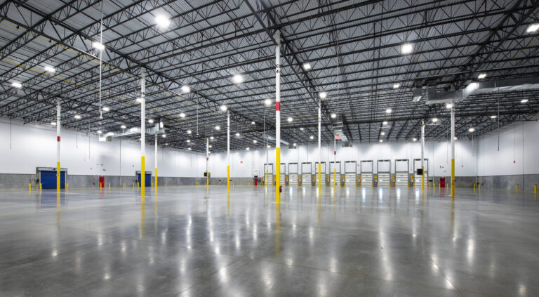 Interior of a large, industrial warehouse with high ceilings, numerous loading docks, polished concrete floors, and extensive lighting.