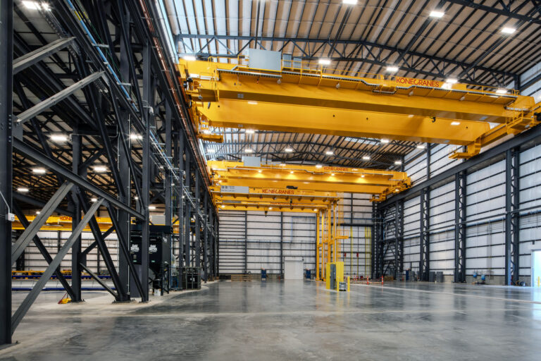 Interior of a large industrial warehouse with high ceilings, featuring two large yellow overhead cranes and multiple industrial steel beams.