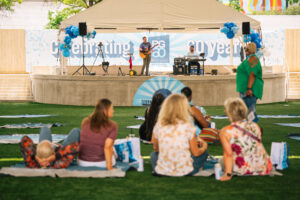 A small crowd sits on a lawn watching a live music performance on an outdoor stage. The stage setup includes musical instruments, speakers, and large banners reading "Celebrating 60 years." An auto draft of the event's schedule appears behind the musicians, adding to the anticipation.