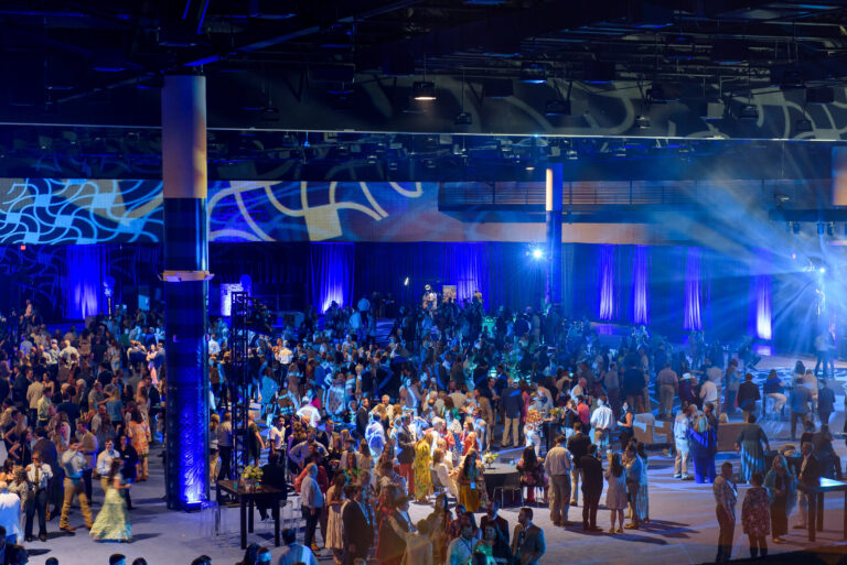 A large crowd attends the Brasfield & Gorrie 60th anniversary party, an indoor event with blue lighting, illuminated stage, and decorative lighting effects on the walls. Various attendees are mingling and talking.