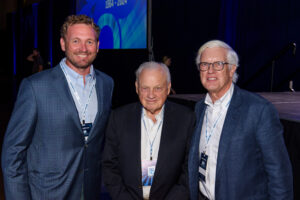 Rob Blalock, Miller Gorrie and Jim Gorrie pose in business attire for a photo at Brasfield & Gorrie's 60th anniversary party, all wearing name badges. The background features a blue banner and stage.