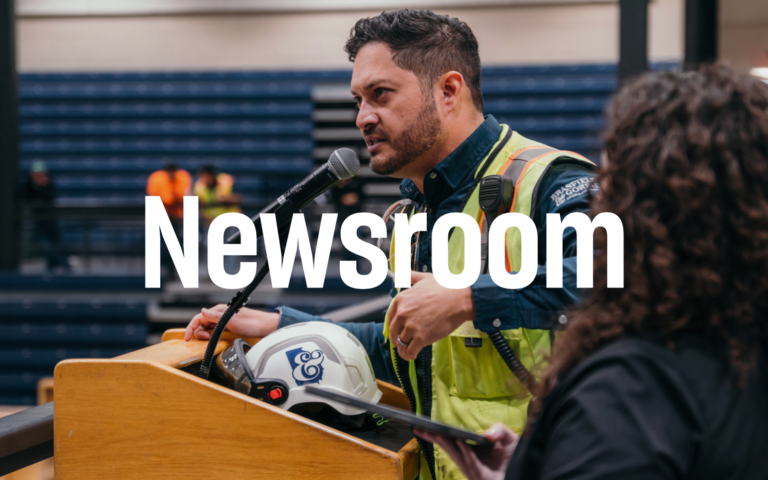 A man in a safety vest speaks into a microphone at a podium. Another person stands beside him holding documents. The word 