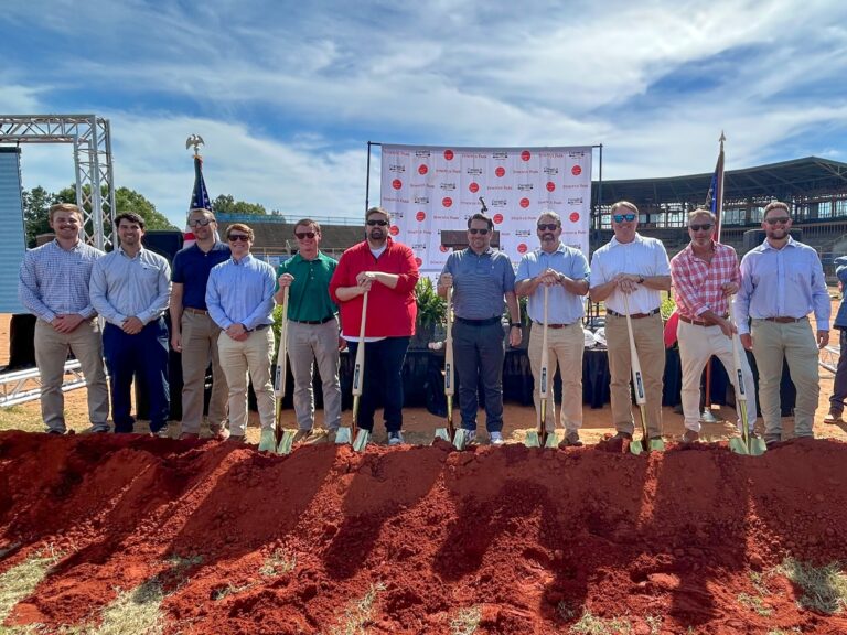 A group of eleven men stand in a line outdoors holding shovels for a groundbreaking ceremony on a red dirt site in Columbus, Georgia. A banner and blue sky are visible in the background.