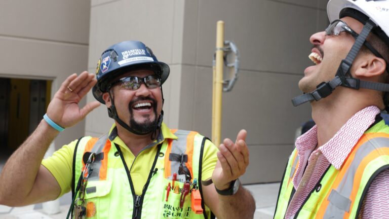 Two construction workers in safety gear are laughing and gesturing while on a jobsite.