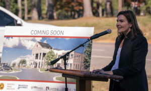 A woman speaks at a podium next to a "Coming Soon" sign featuring a building rendering.