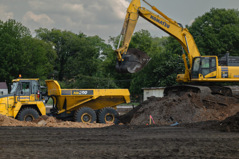 At a self-perform construction site surrounded by trees, a yellow excavator efficiently loads dirt into a matching dump truck.