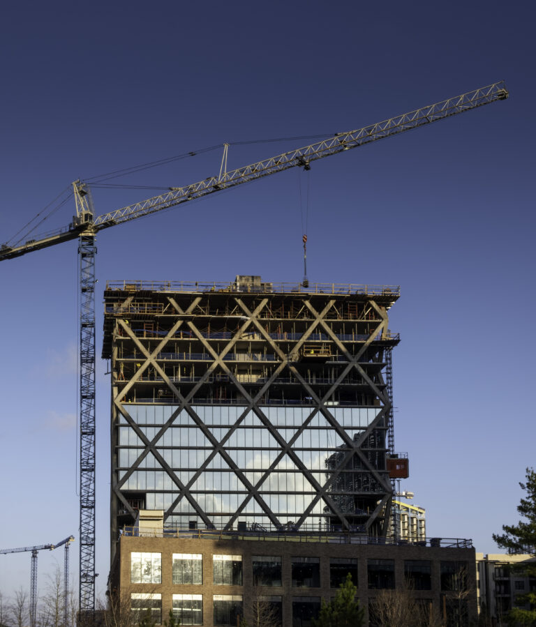 High-rise building under self-perform construction with a diagonal grid pattern facade. A crane is positioned at the top, and the sky is clear.