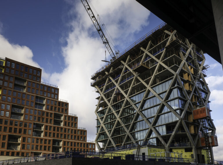 A construction site by Brasfield & Gorrie features a modern building with a striking geometric lattice design and a crane overhead, adjacent to a completed brown brick building under a cloudy sky.