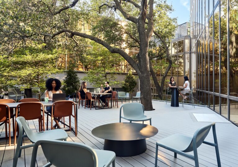 The outdoor patio boasts a thoughtful design, with people sitting at tables surrounded by trees. Cushioned chairs and tables are arranged on a wooden deck, harmonizing with the glass building facade in the background.