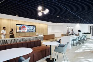 A modern office lounge features round tables, chairs, and a TV on the wall. Amid bright lighting and a blue ceiling, people engage in conversation, showcasing a space where design meets functionality.