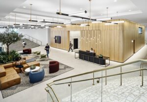 The modern office lobby, with its sleek design, features a wooden reception desk under stylish lighting fixtures. People mingle around the seating area surrounded by lush plants and elegant stairs.