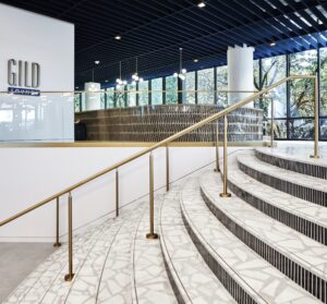 A masterful design graces the modern lobby, where a curved staircase with brass railings elegantly leads to a repositioned lounge area. Large windows flood the space with natural light, while a sign reads "Guild Lounge.